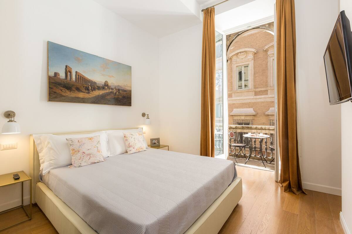 A hotel room with a bed, a painting on the wall, a TV, and a balcony with a table and chairs.