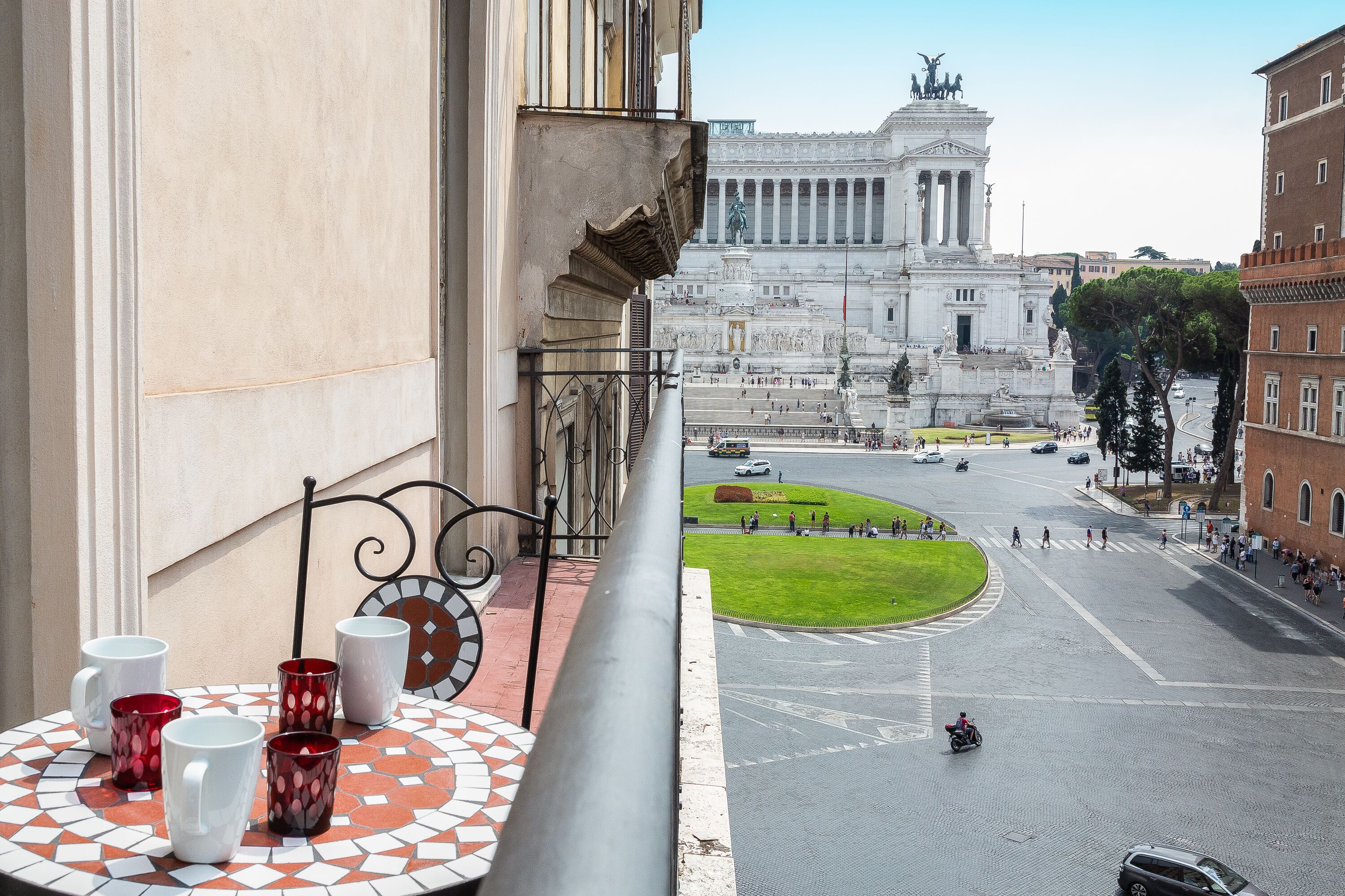 A balcony with a tiled table and four mugs, overlooking a city square with a large monument and a roundabout.