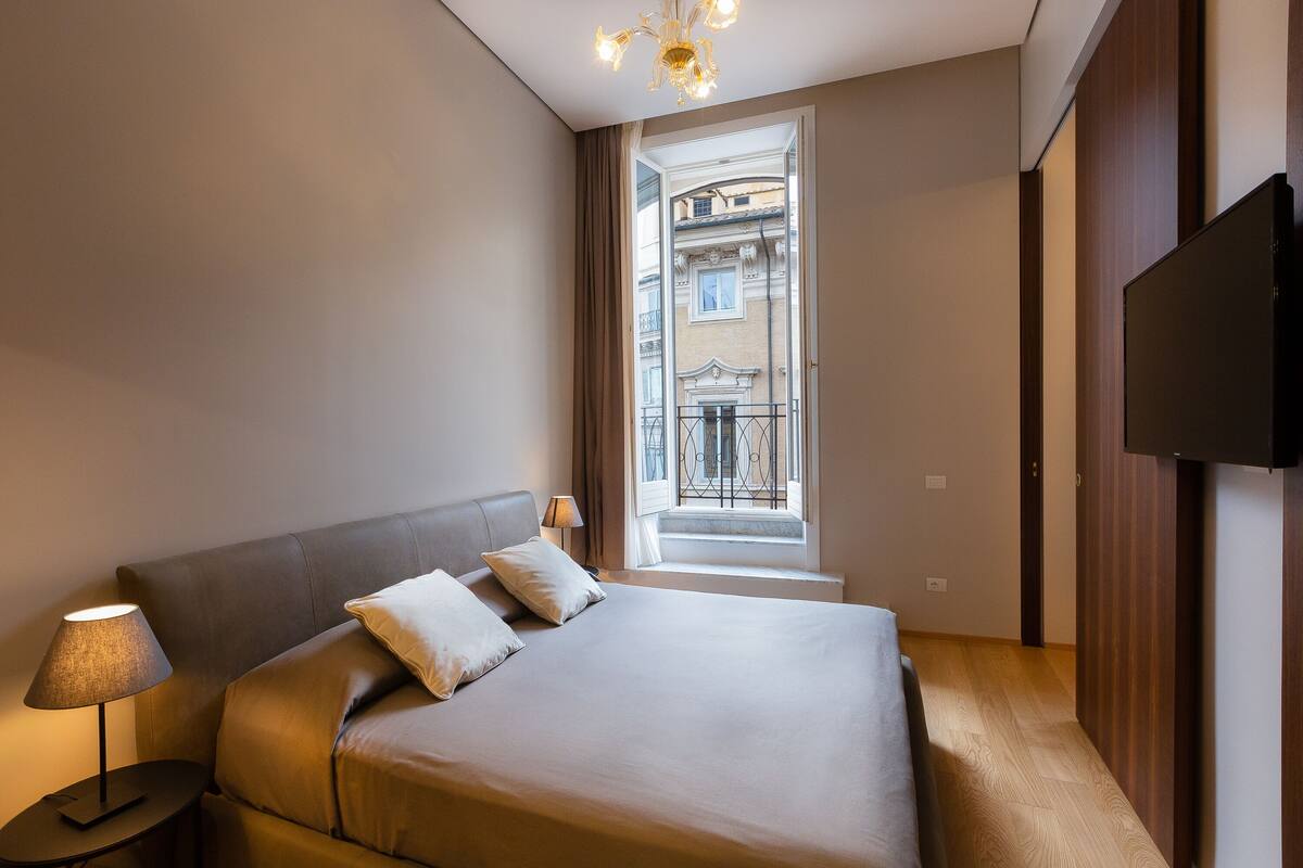 A hotel room with a large bed, a bedside table with a lamp, a TV mounted on the wall, and a view of a building through a window.