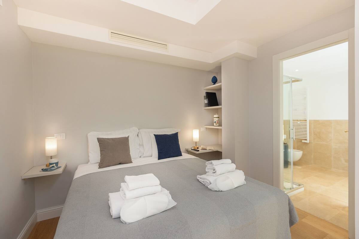 A hotel room with a bed, bedside tables, a nightstand, a lamp, a shelf with decorative items, and a view of the bathroom.
