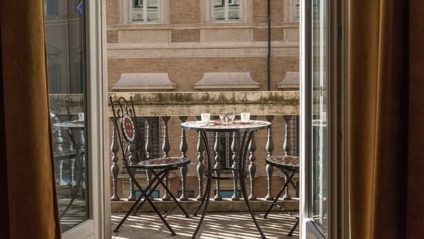 Een balkon buiten met twee ronde tafels en stoelen, uitzicht op een bakstenen gebouw met ramen, en een hekwerk.
