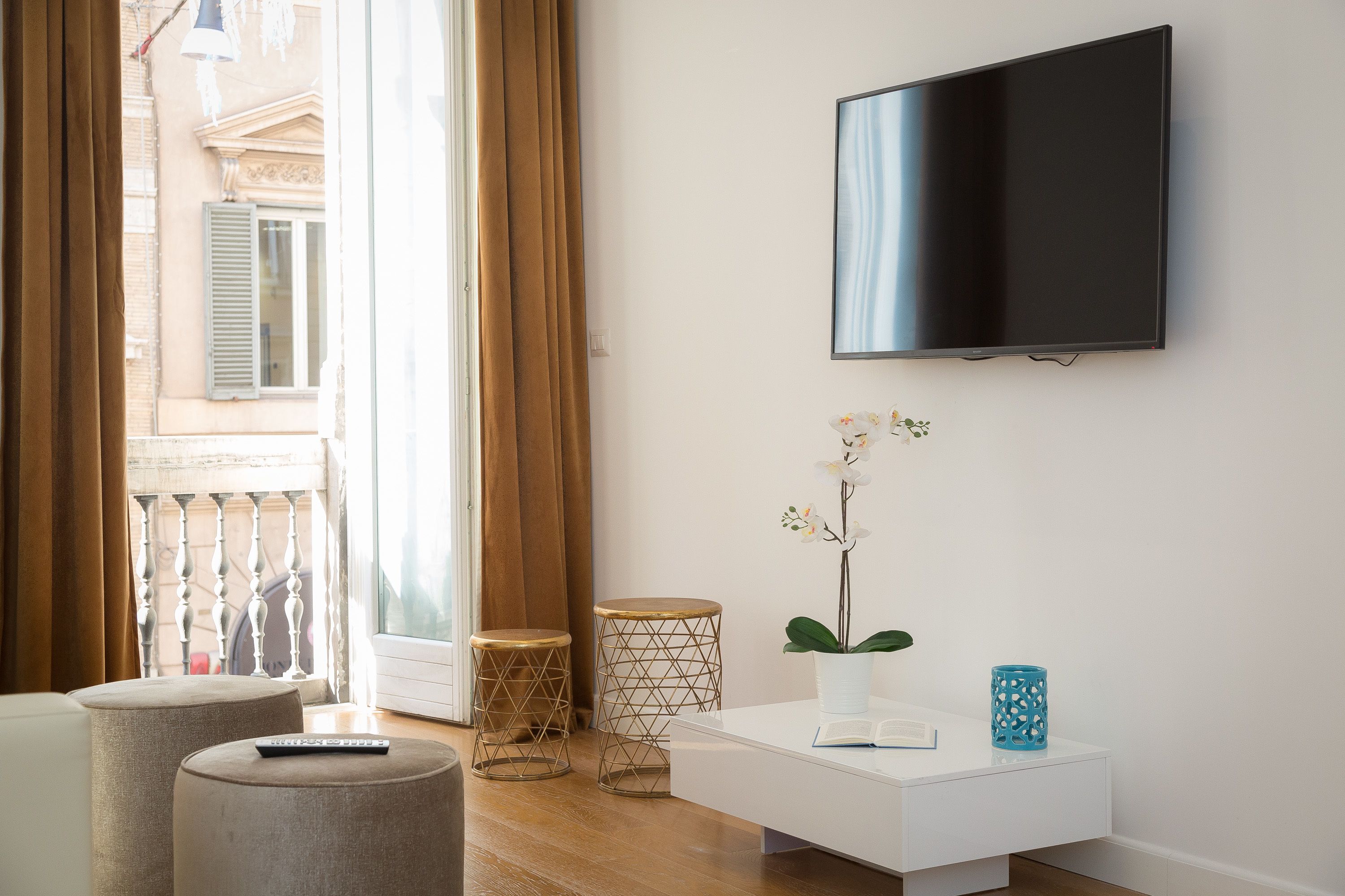 Ett modernt vardagsrum med en platt-TV, ett vitt soffbord och en orkidé i en kruka. Det finns två runda pallar och ett litet runt bord med ett vävt mönster.