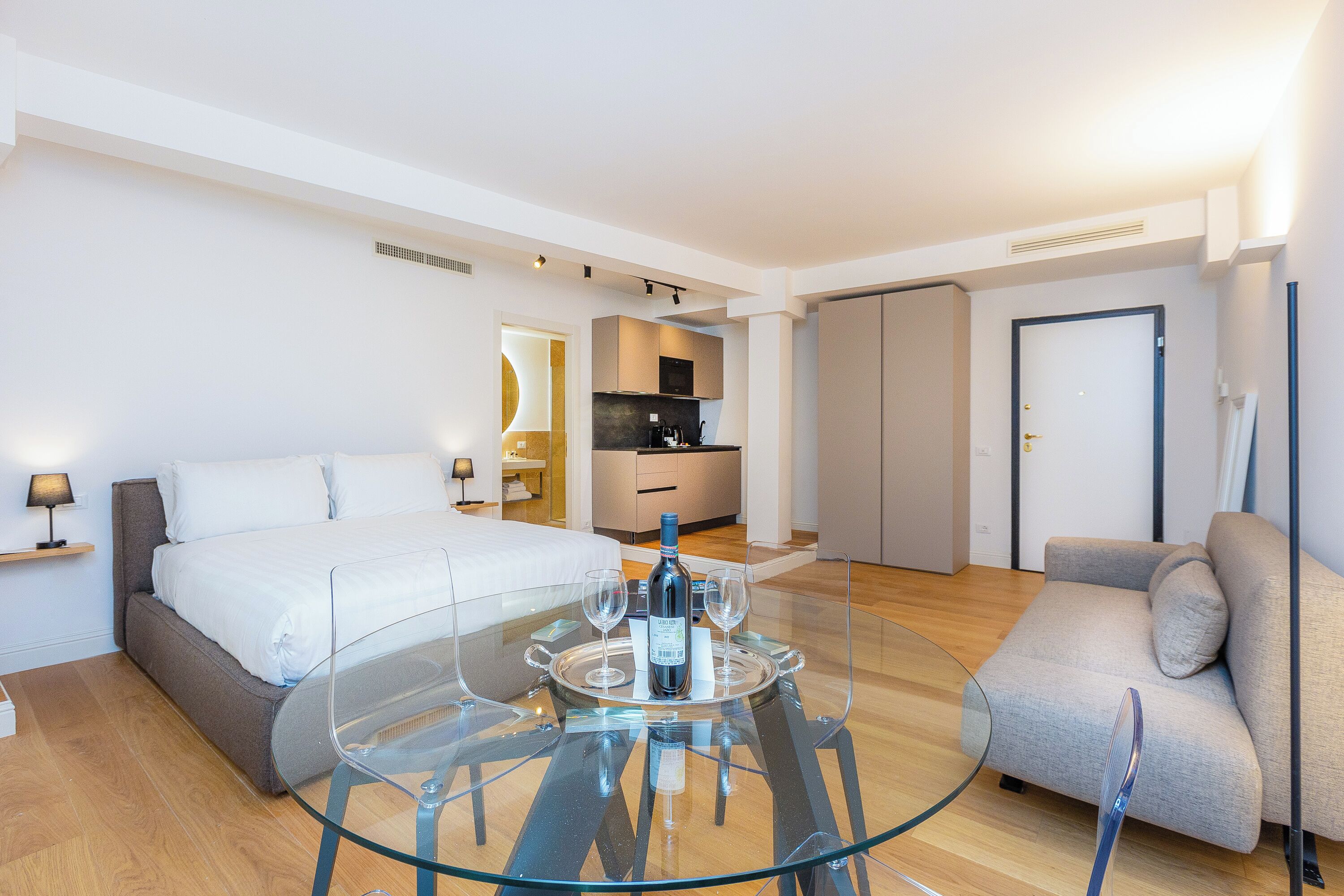 Una moderna camera d'hotel con un letto, un tavolo da pranzo in vetro con bicchieri da vino, una poltrona e, sullo sfondo, un angolo cottura.