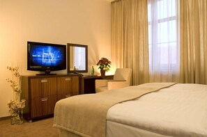 A hotel room with a bed, a TV, a desk with a chair, a plant, and a window with curtains. - Hotel Academic (Zvolen)