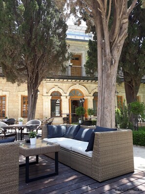 An outdoor seating area with wicker furniture and a glass table, surrounded by trees and a building with arched windows. - Sergei Palace Hotel (Jerusalem)