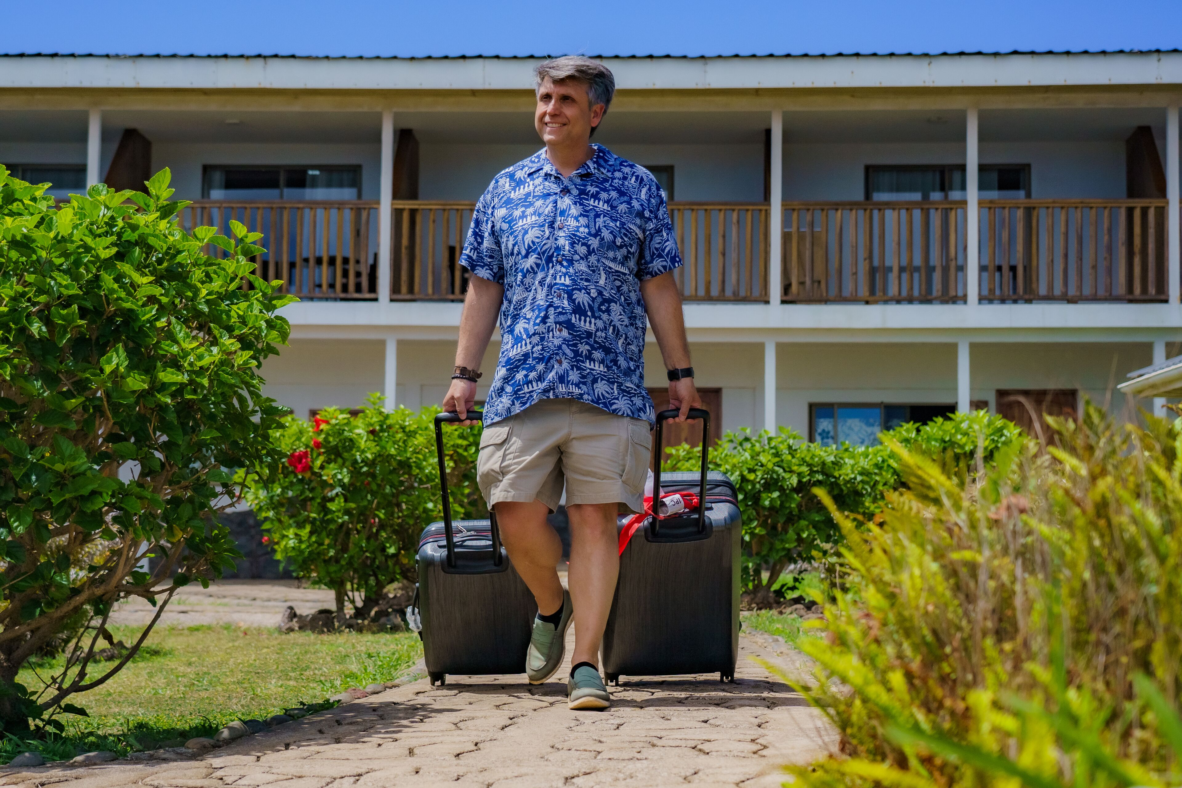 A man walking with two suitcases on a paved path in front of a building with balconies.