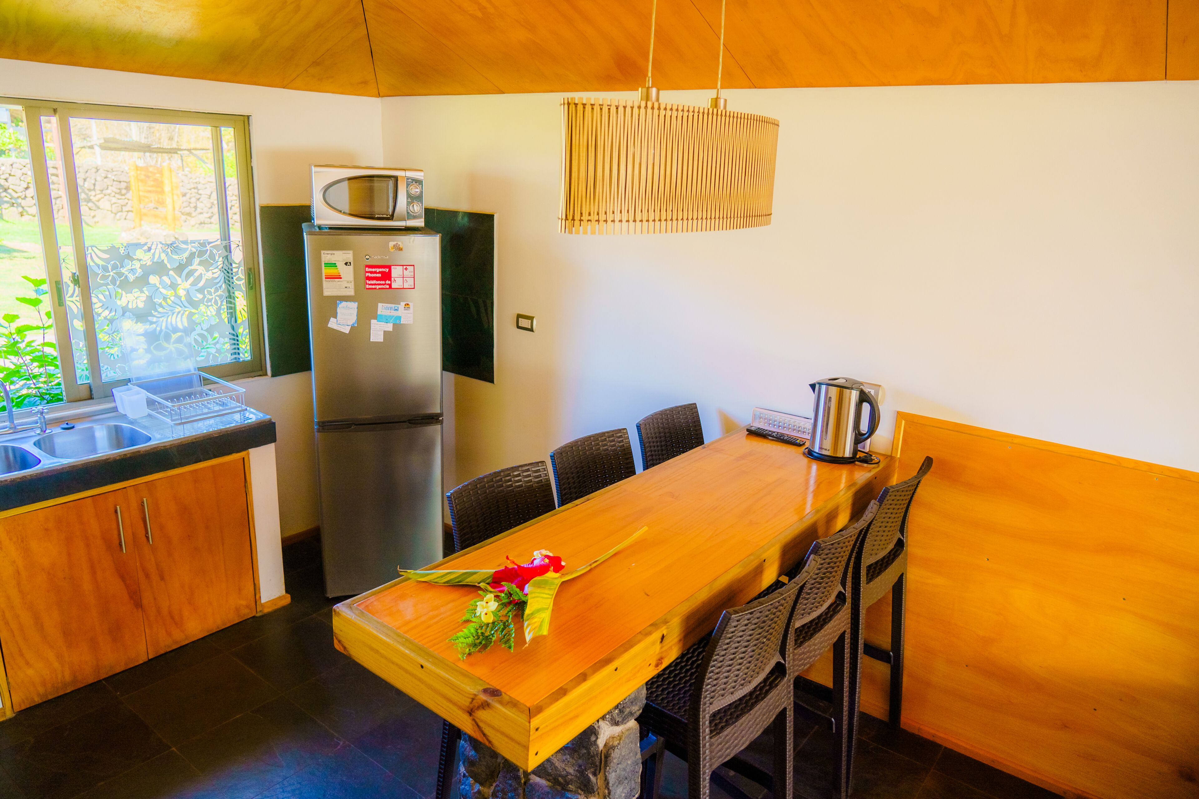 A compact kitchen with a wooden table, dark wicker chairs, a refrigerator, a microwave, and a hanging light fixture.