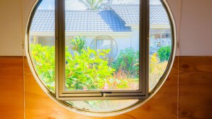 Een rond raam met uitzicht op een huis met pannendak en omliggend groen.