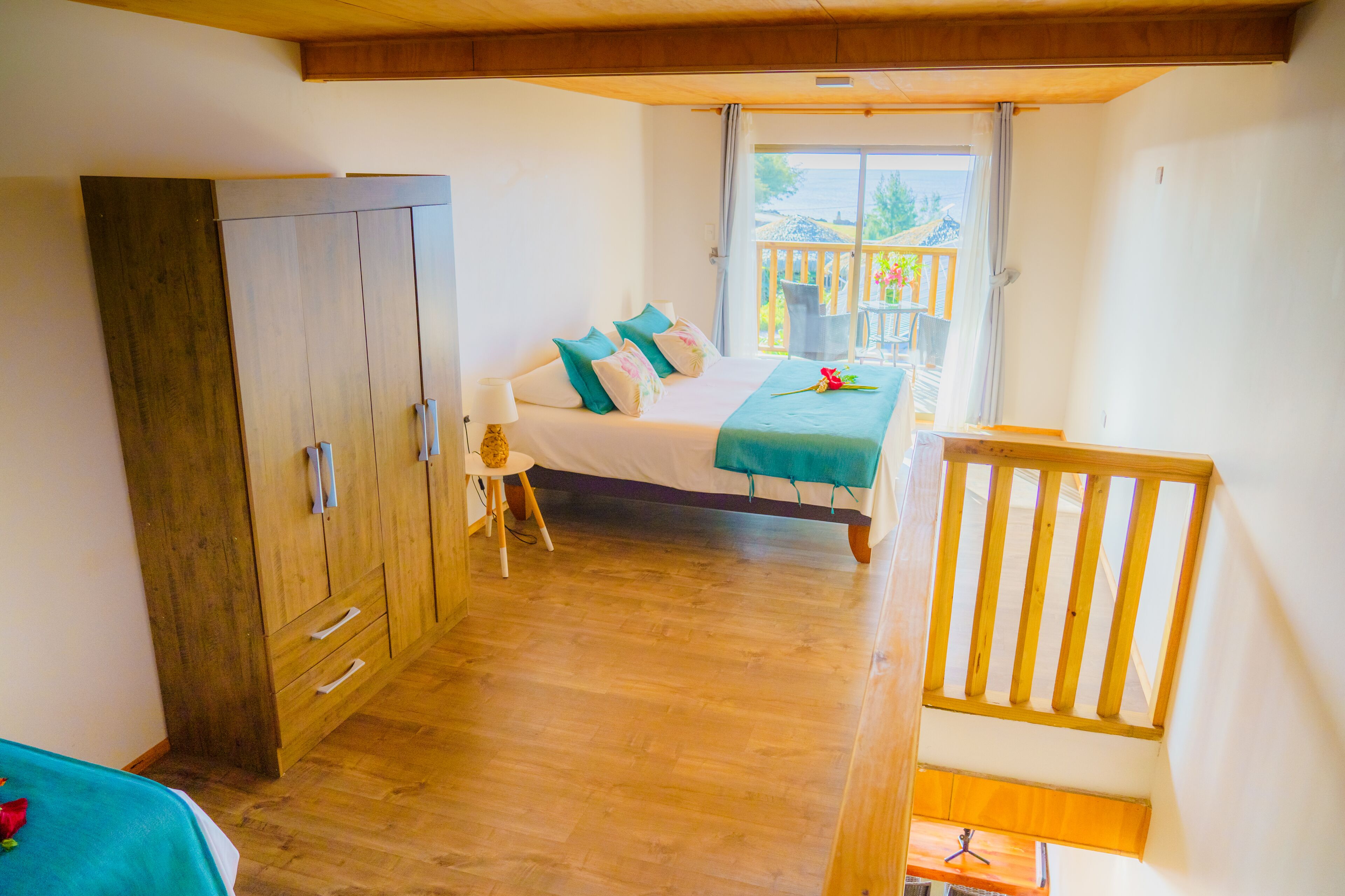 A bedroom with a bed, a wooden wardrobe, a small table with a lamp, and a balcony with a railing.