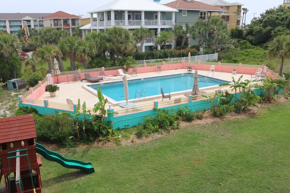A swimming pool surrounded by a deck with lounge chairs and a slide, set against a backdrop of multi-story buildings and lush greenery. - Deep Water Cove by Surfside VR (Pensacola Beach)