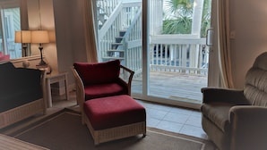 A living room with a red cushioned chair, a wicker chair, a small table with a lamp, and a view of a wooden deck and stairs leading outside. - Boardwalk by Surfside VR (Pensacola Beach)