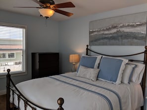 A bedroom with a bed, a ceiling fan, a window, a lamp, and a painting on the wall.