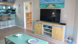 A living room with a TV, a dining table, and a kitchen area with bar stools. - Sand Dollar by Surfside VR (Pensacola Beach)