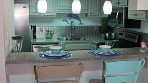 A kitchen with a 'BEACH CAFE' sign, two barstools, a refrigerator, a microwave, and a sink. - Sand Dollar by Surfside VR (Pensacola Beach)
