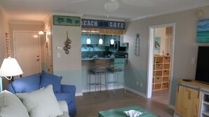 A living room with a blue sofa, a green coffee table, and a kitchen area with a 'BEACH CAFE' sign. - Sand Dollar by Surfside VR (Pensacola Beach)