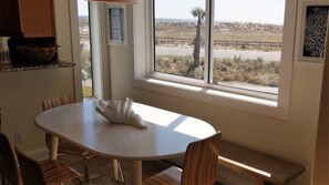 A dining area with a white table and chairs, a window with a view of a beach and palm tree, and a shelf with kitchen items.