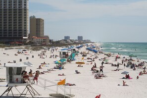 A crowded beach with people sunbathing and swimming, a lifeguard tower, and tall buildings in the background.
