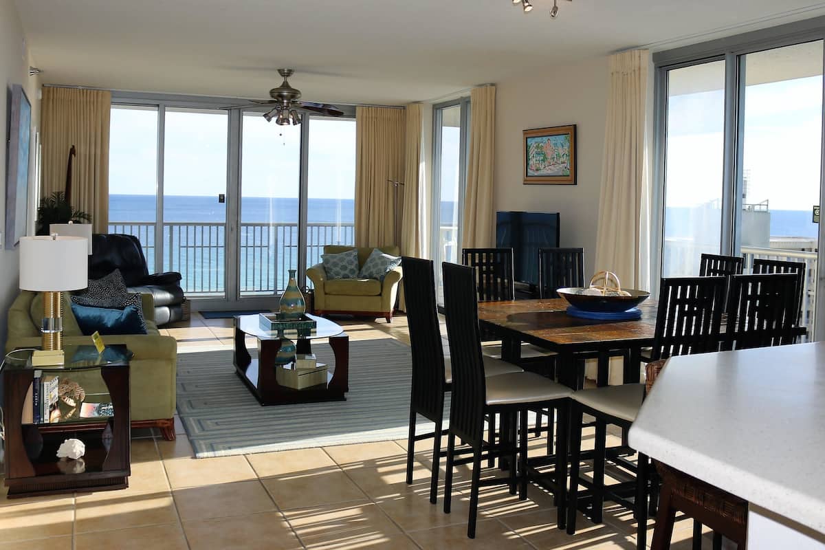 En rummelig opholdsstue med balkon med udsigt over havet, møbleret med sofa, lænestole, spisebord og stole.
