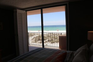 Una habitación con cama, ventana con vista a la playa y al océano, y balcón con barandilla.