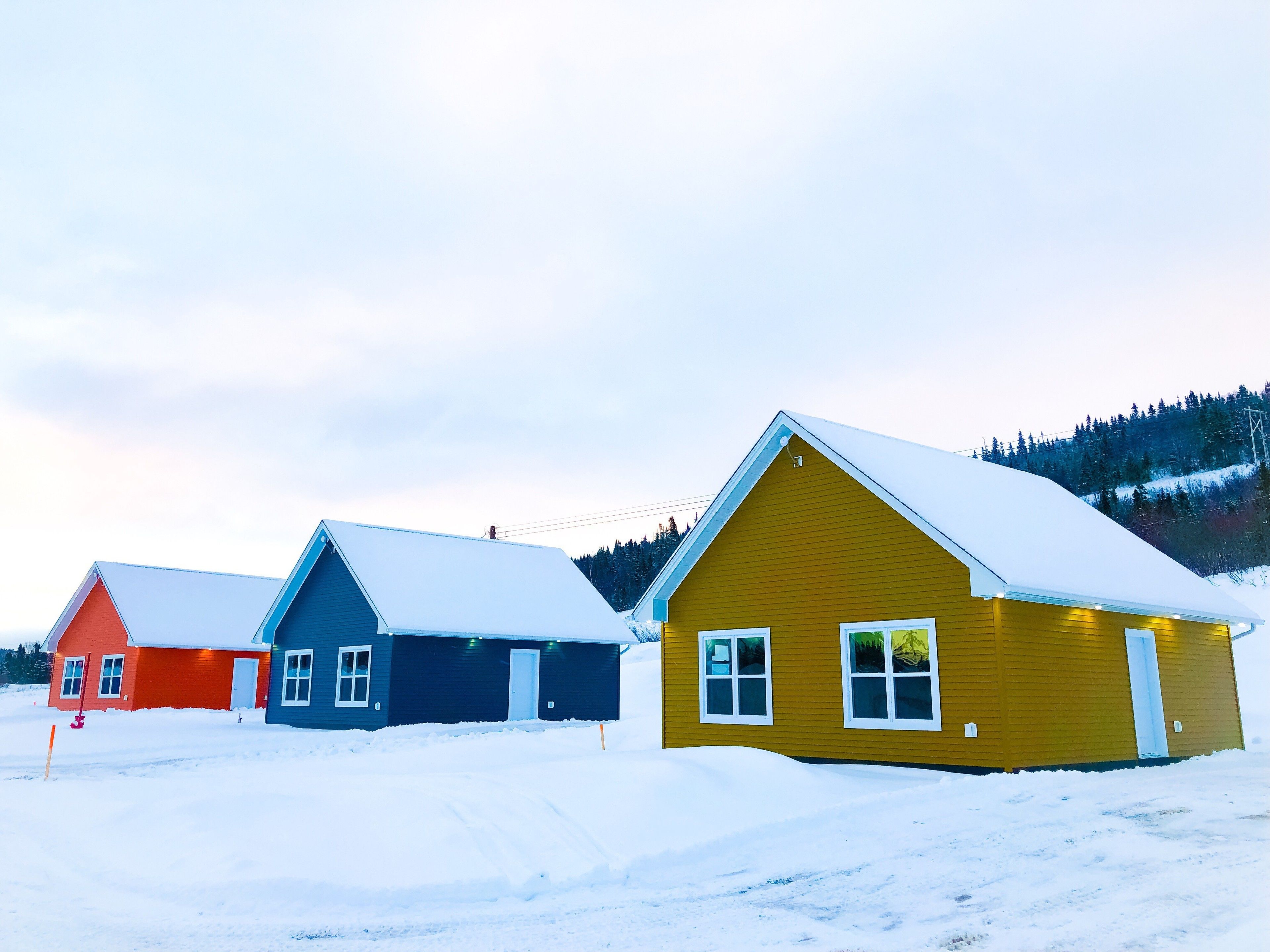 Tres casas alineadas con tejados nevados, con un paisaje invernal y una colina boscosa al fondo.