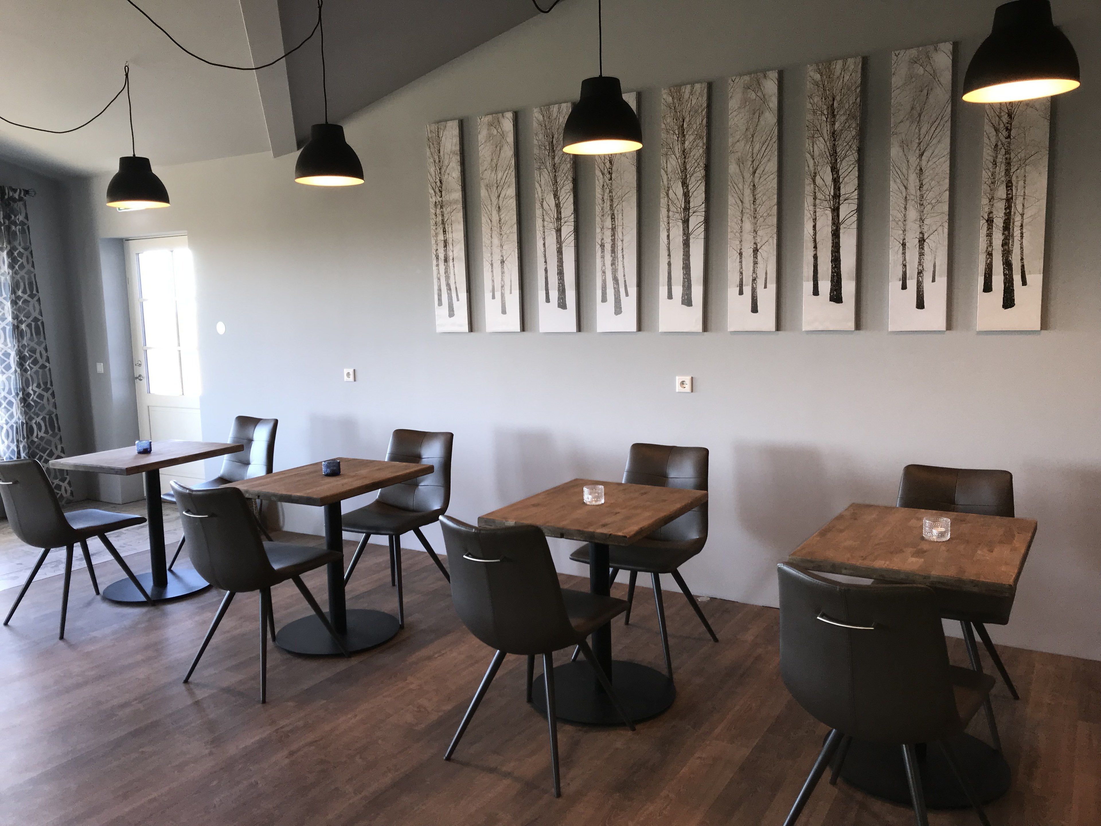 Uma área de refeições moderna com mesas de madeira e cadeiras pretas, apresentando uma fileira de pinturas abstratas de árvores na parede.