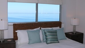 A hotel room with a large window offering a view of the sea, a bed with white linens and two pillows, a patterned headboard, and two bedside lamps. - Tristan Towers by Surfside VR (Pensacola Beach)