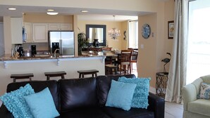 A modern kitchen with a refrigerator, coffee maker, and dining area with chairs and a table. - Tristan Towers by Surfside VR (Pensacola Beach)