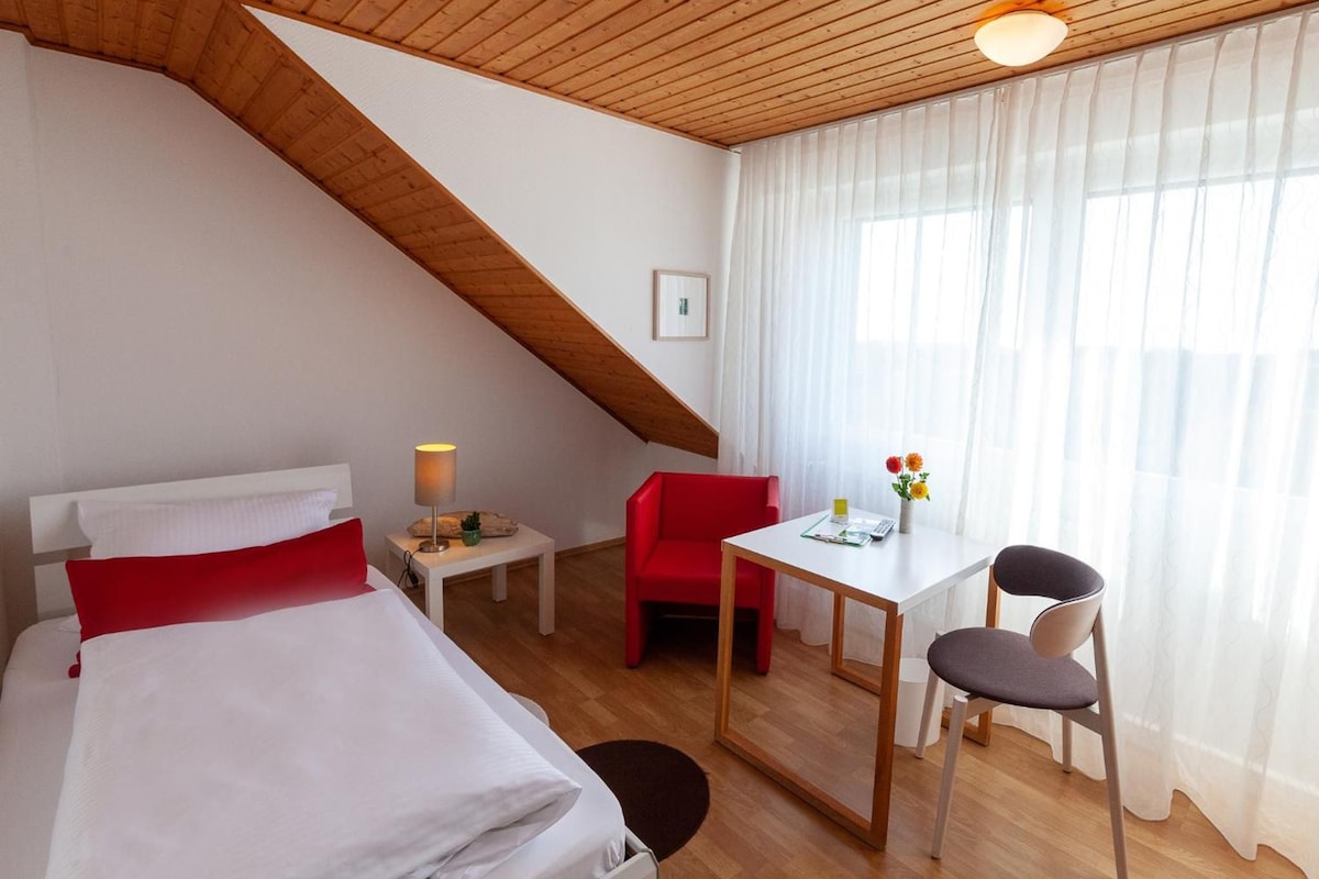 A hotel room with a sloped ceiling, a bed with white bedding and red pillows, a small desk with a chair, a red chair, and a window with sheer curtains.