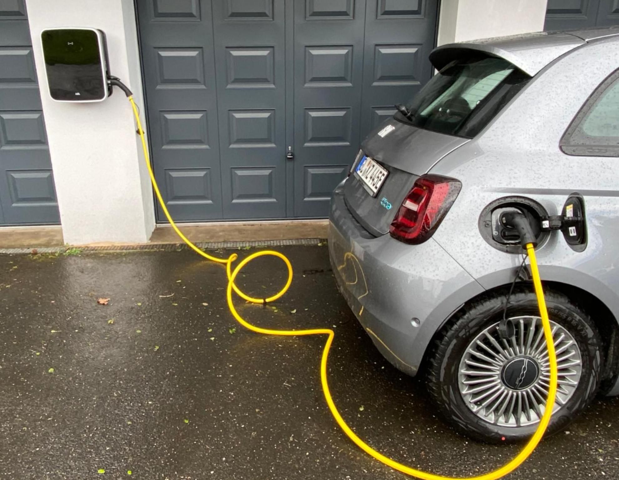 Ein silbernes Auto steht vor einer grau lackierten Doppelgarage, ein gelbes Ladekabel ist an der Lademulde des Autos angeschlossen.