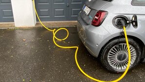 A silver car parked in front of a grey double door garage with a yellow charging cable connected to the car's charging port.