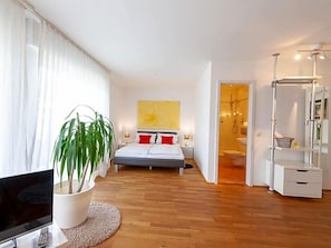 A modern bedroom with a bed, bedside tables, a TV, a potted plant, and a bathroom visible through an open door.