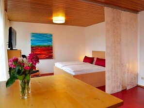 Una habitación con cama, un cuadro, una mesa con flores y una cortina.