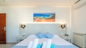 A hotel room with a bed, two bedside tables, a nightstand, a painting of a coastal landscape, and a view of the sea. - Hotel Martorell (Ses Salines)