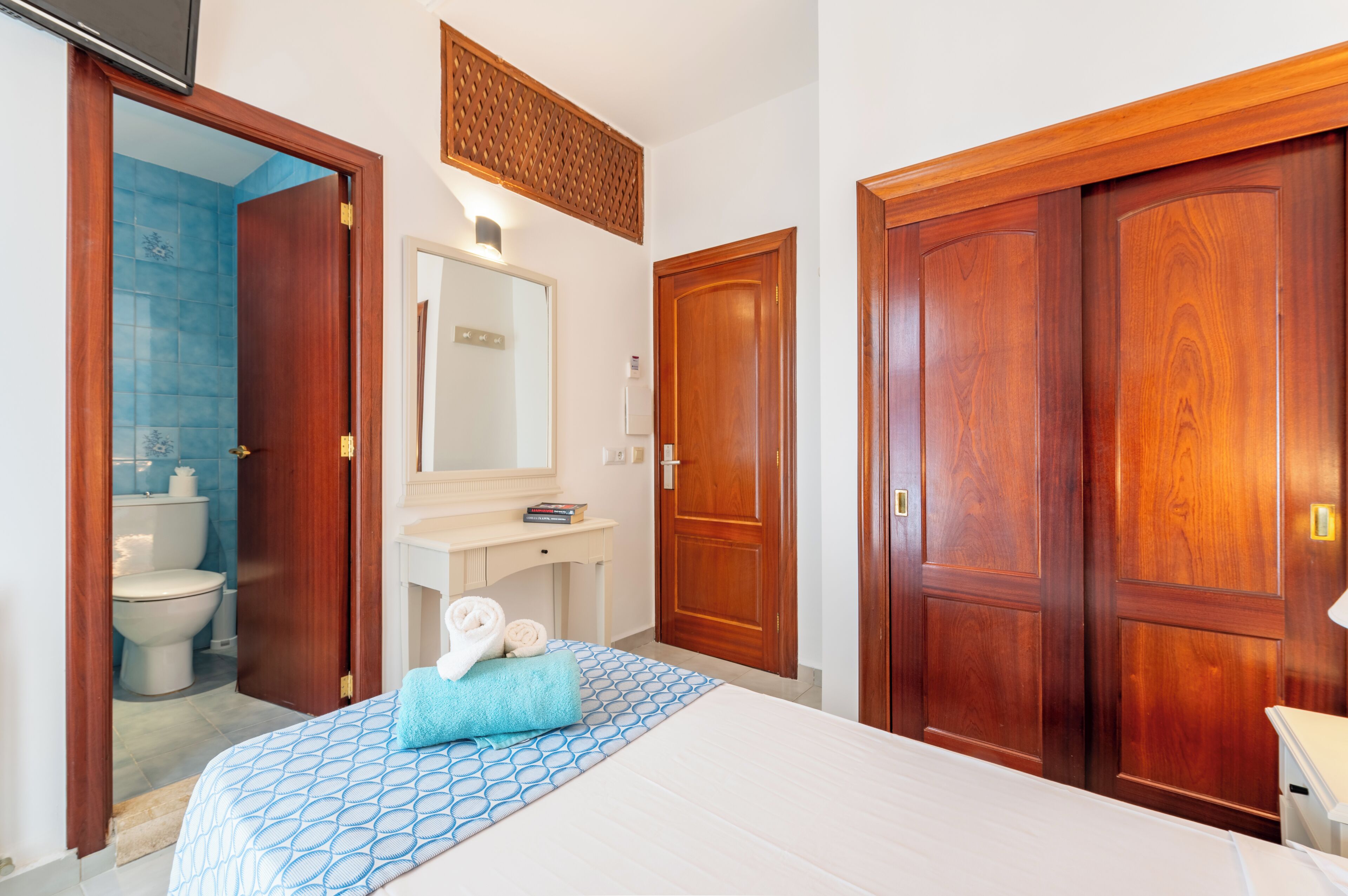 Quarto de hotel com uma cama, banheiro com vaso sanitário e pia, e portas de madeira.