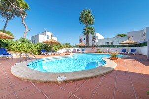Zona de piscina con sillas de descanso y sombrillas, rodeada de edificios blancos y vegetación.