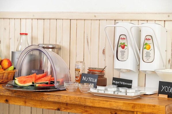 A breakfast bar with a fruit display, coffee machines, and condiments. - Hotel Martorell (Ses Salines)