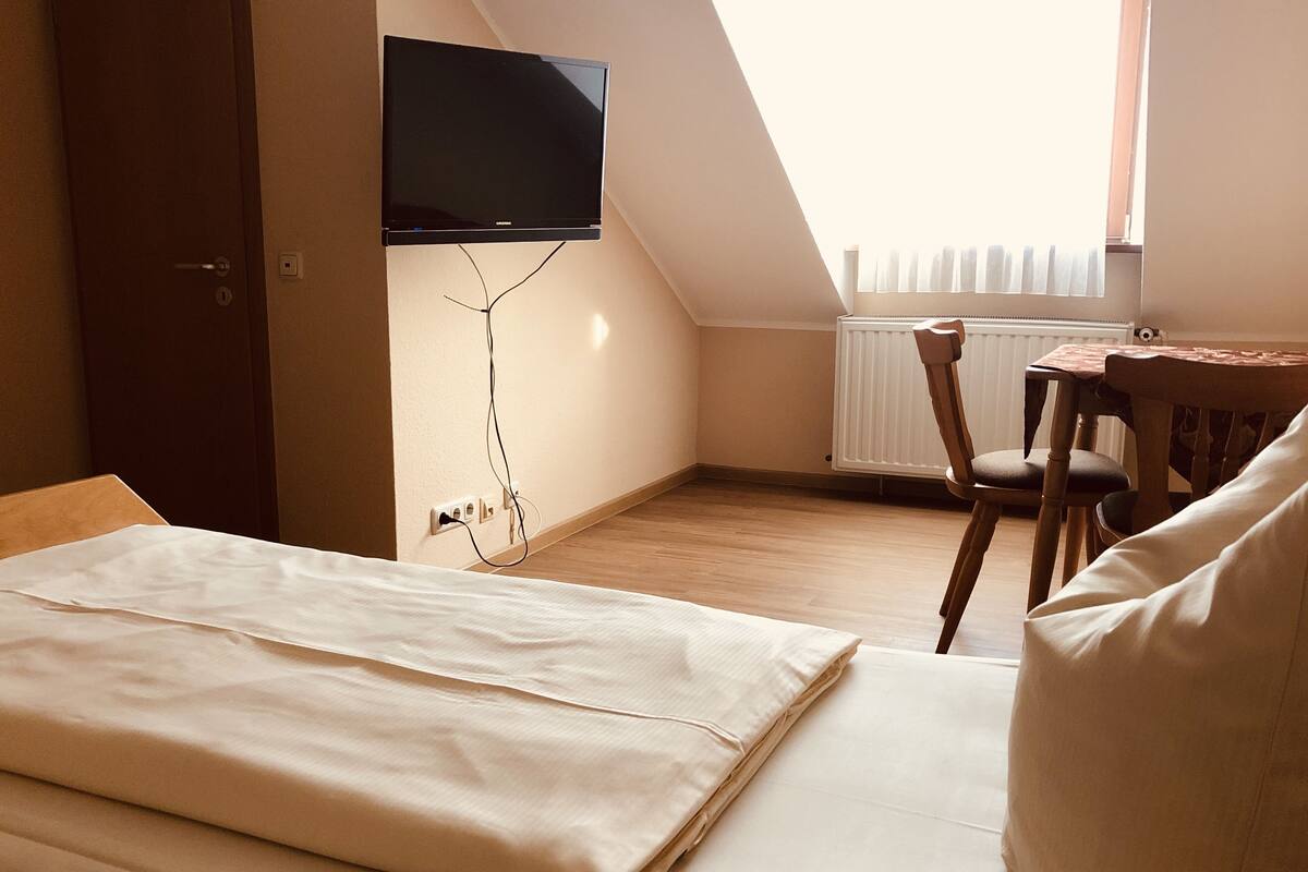 A bedroom with a bed, a TV mounted on the wall, a wooden table, and a chair.