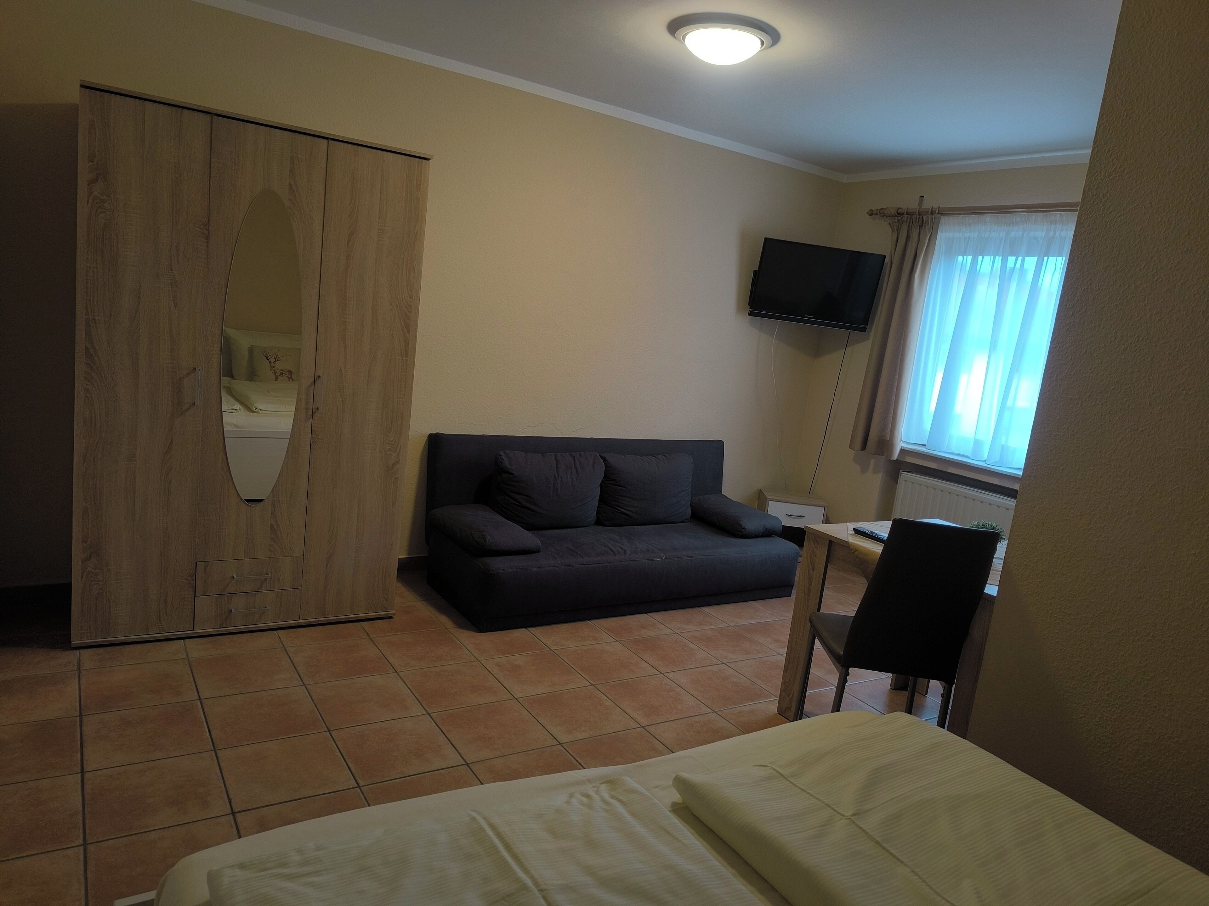 A bedroom with a bed, a dark sofa, a wooden wardrobe, a television, and a window with curtains.