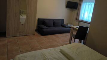 A bedroom with a bed, a dark sofa, a wooden wardrobe, a television, and a window with curtains.