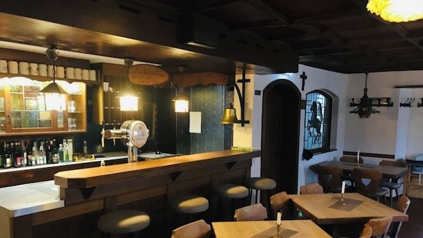 Una zona de bar con mesas y taburetes de madera, una barra con botellas y luces colgantes.