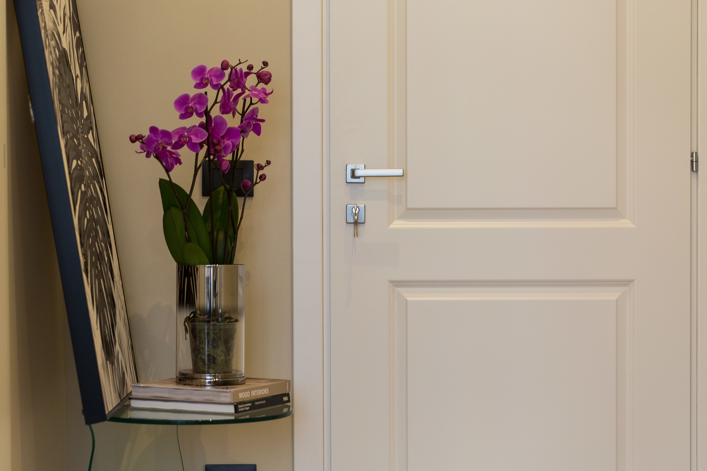 A door with a keyhole and a handle, a glass shelf with a book titled "WOOD INTERIORS," and a vase with purple flowers.