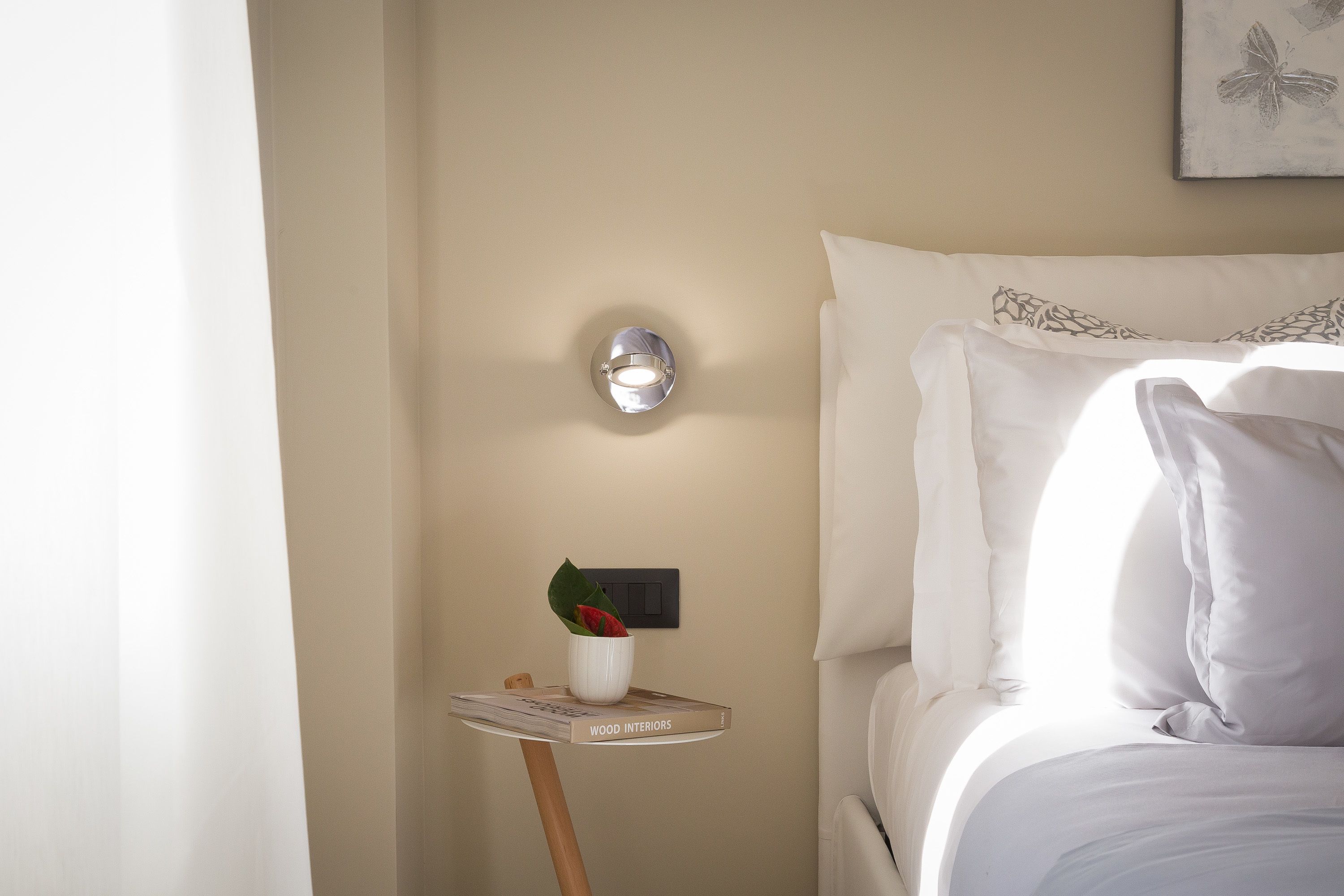 A neatly made bed with white linens and a small table beside it holding a book titled "WOOD INTERIORS" and a white vase with a red flower.