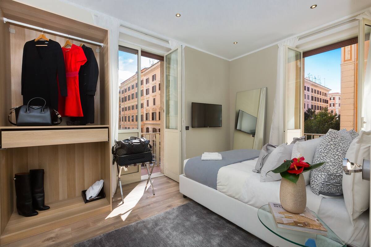 A modern hotel room with a bed, a TV, a wardrobe, and a view of buildings.