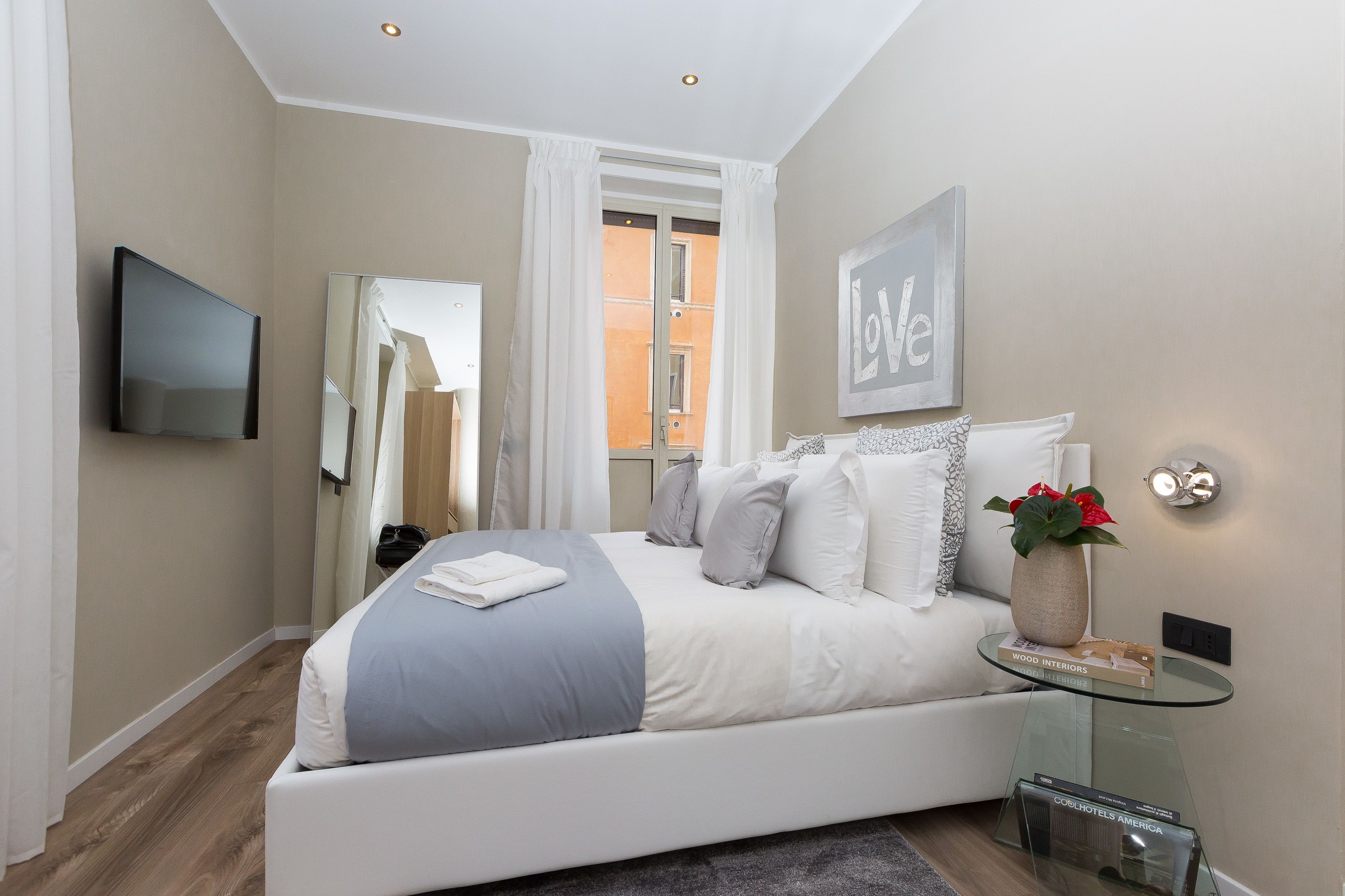 Un dormitorio moderno con una cama, una mesita de vidrio, un televisor montado en la pared y un ventanal con cortinas.
