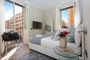 A modern hotel room with a bed, a glass table, a TV, and a view of buildings. - 126 Gracchi Suites (Rome)