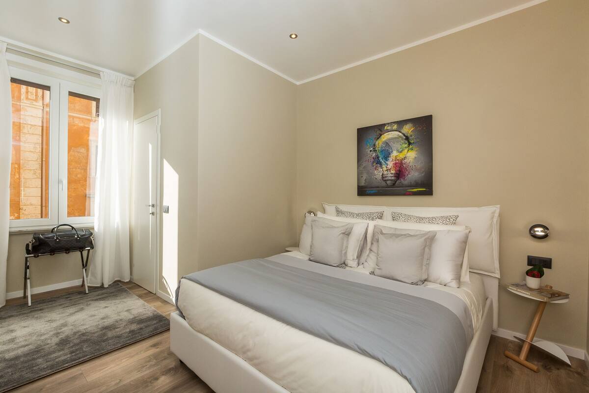 A bedroom with a bed, a bedside table, a wall art piece, and a window with curtains.