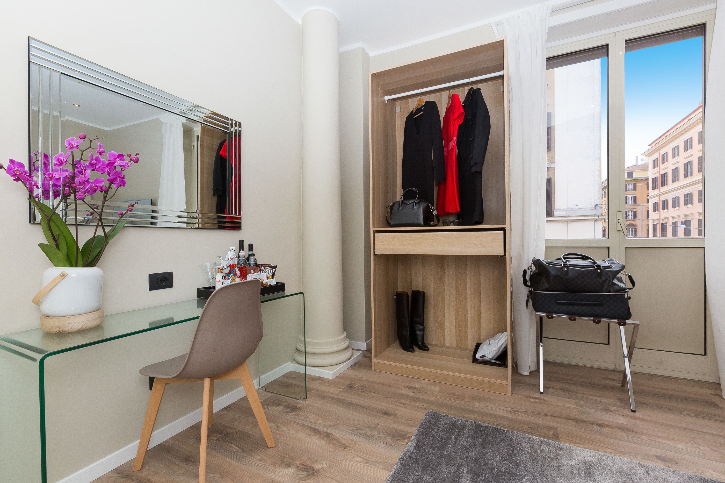 Ein modernes Zimmer mit einem Glastisch, einem Stuhl, einem Spiegel, einem Kleiderschrank und einem Fenster mit Blick auf Gebäude.