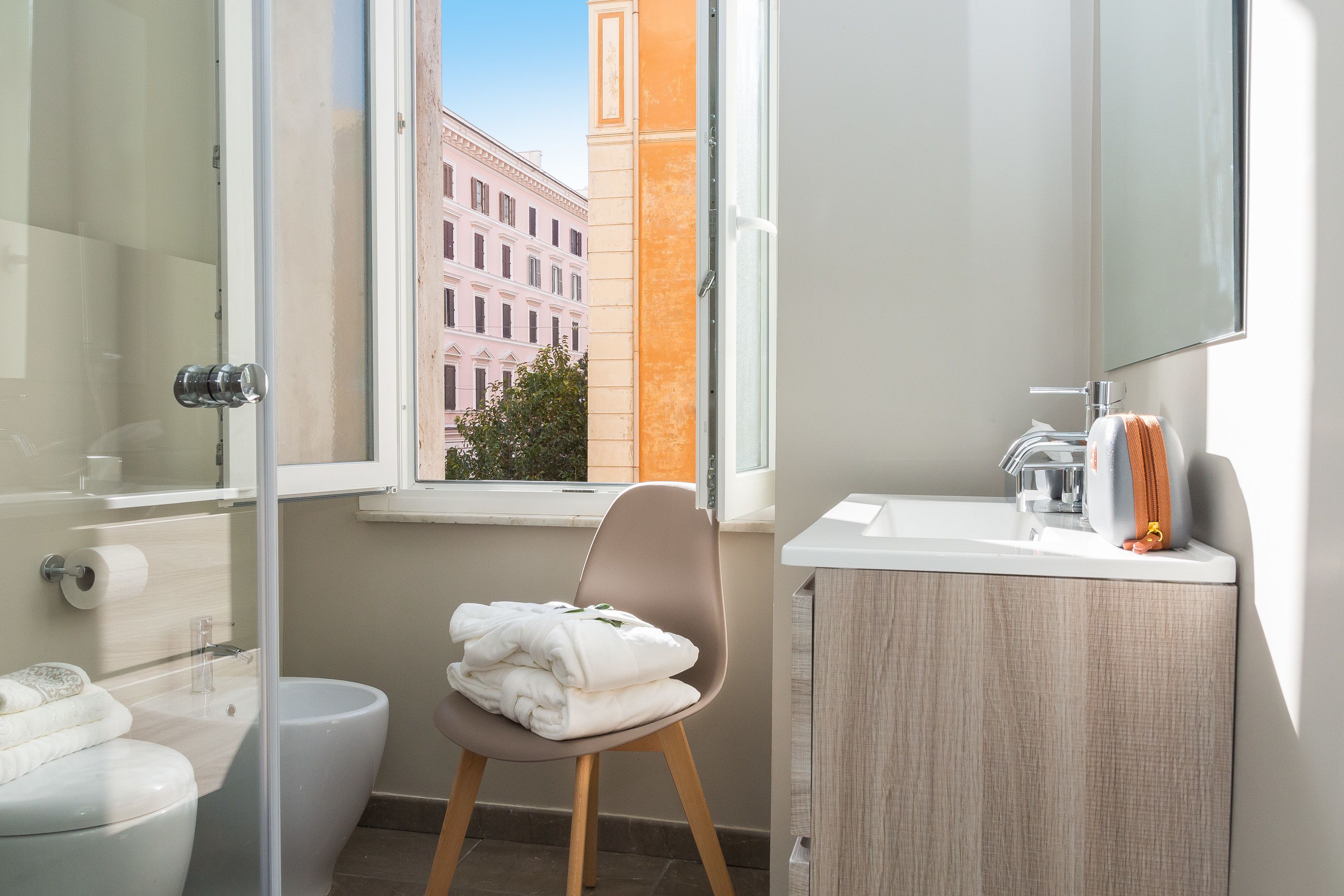 A modern bathroom with a glass shower enclosure, a toilet, a sink with a mirror, and a window with a view of a building.