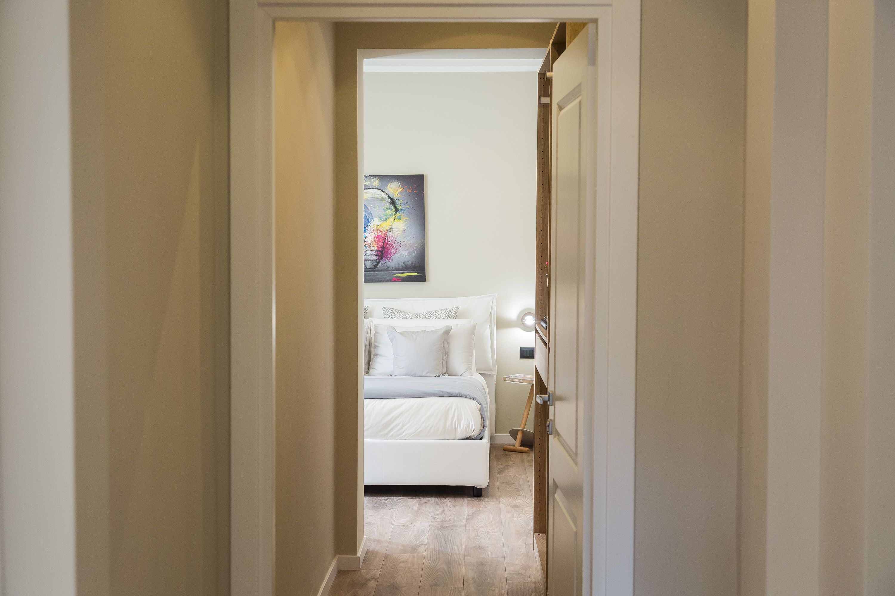 Un pasillo estrecho que conduce a un dormitorio con cama, mesita de noche y aplique en la pared.