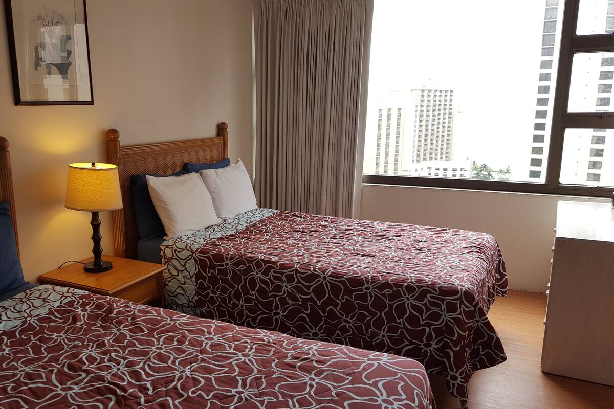 Habitación de hotel con dos camas, una mesita de noche con lámpara, un cuadro en la pared y una ventana con vistas a edificios.